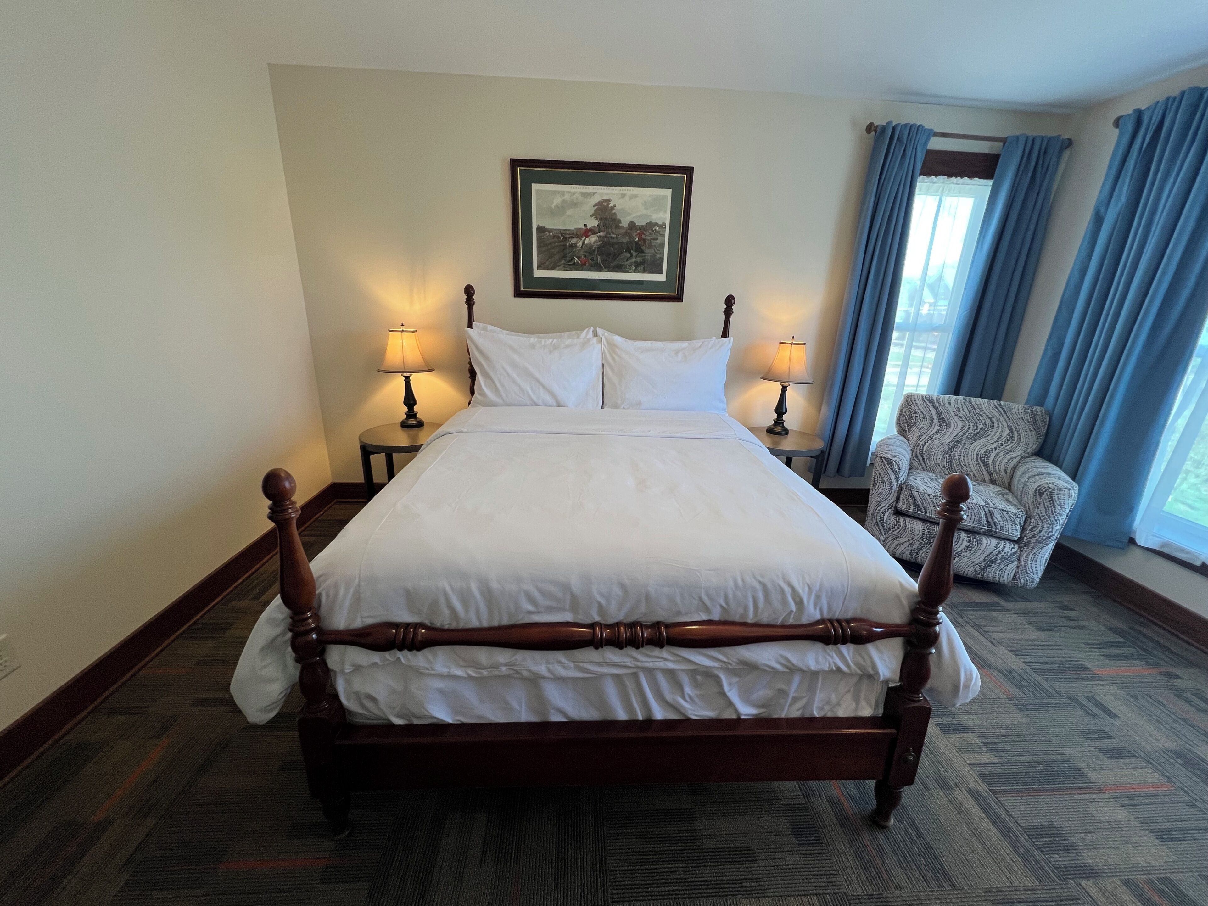 A hotel room with a large bed, two bedside tables with lamps, a chair, a framed picture on the wall, and a window with curtains.