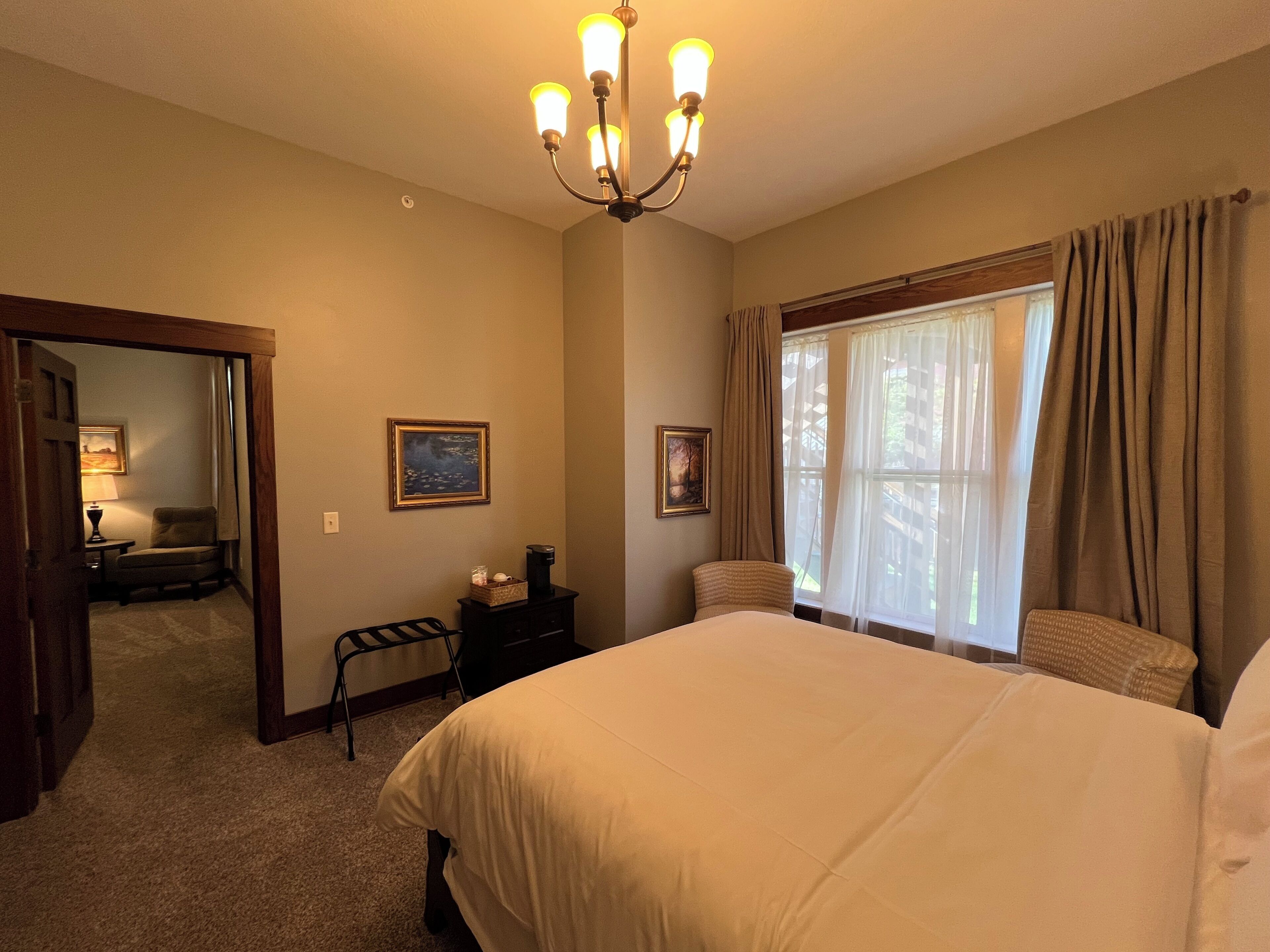 A hotel room with a large bed, a desk with a lamp, a chair, a coffee maker, and a painting on the wall.