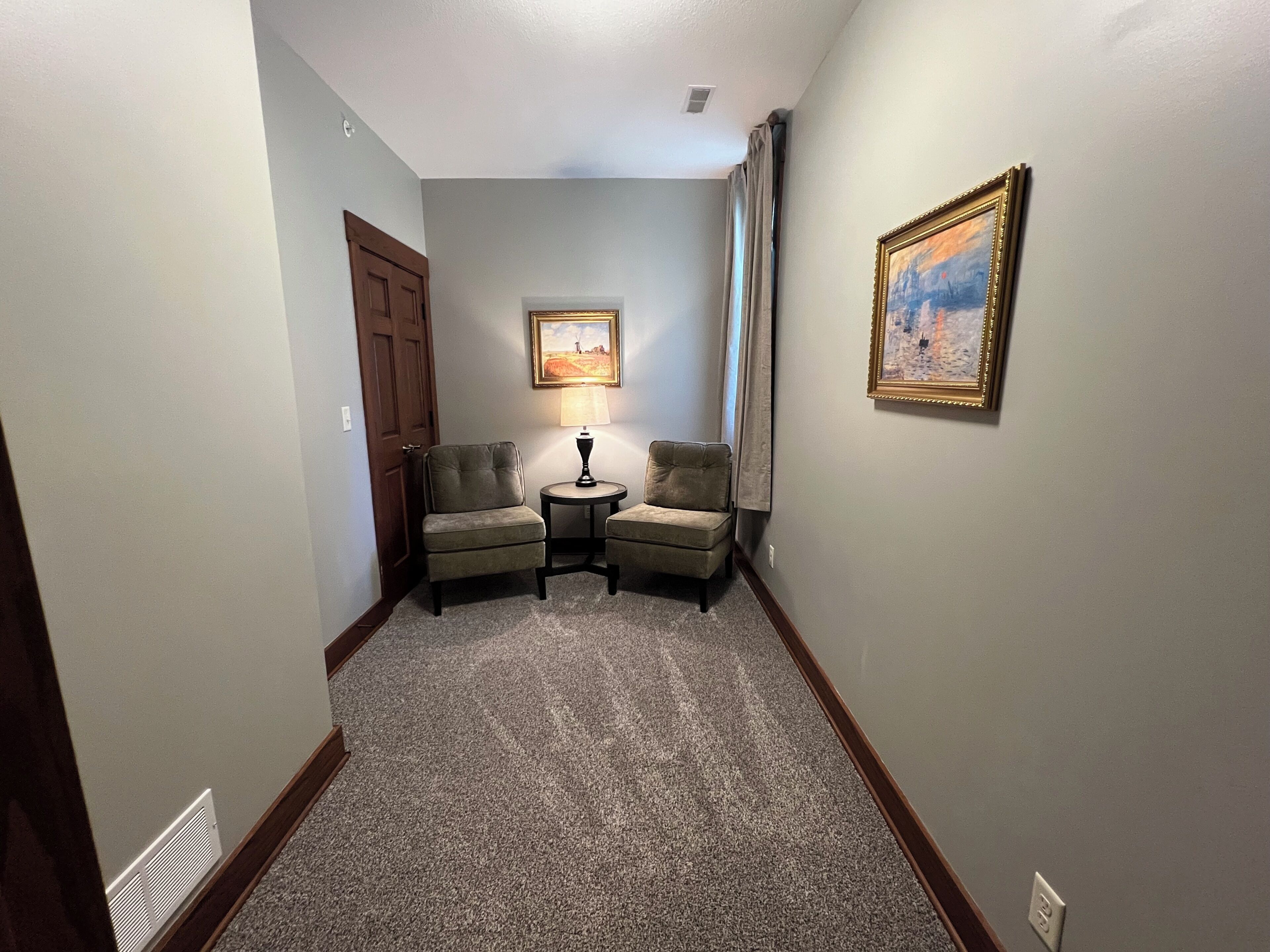 Un pasillo con piso alfombrado, dos sillones, una mesita con lámpara, una puerta y un cuadro en la pared.
