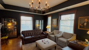 A living room with a brown leather sofa, a beige armchair, and a matching ottoman. There are two side tables with lamps, a chandelier, and a framed portrait on the wall. - Charleston House Bed & Breakfast Inn (Charleston)