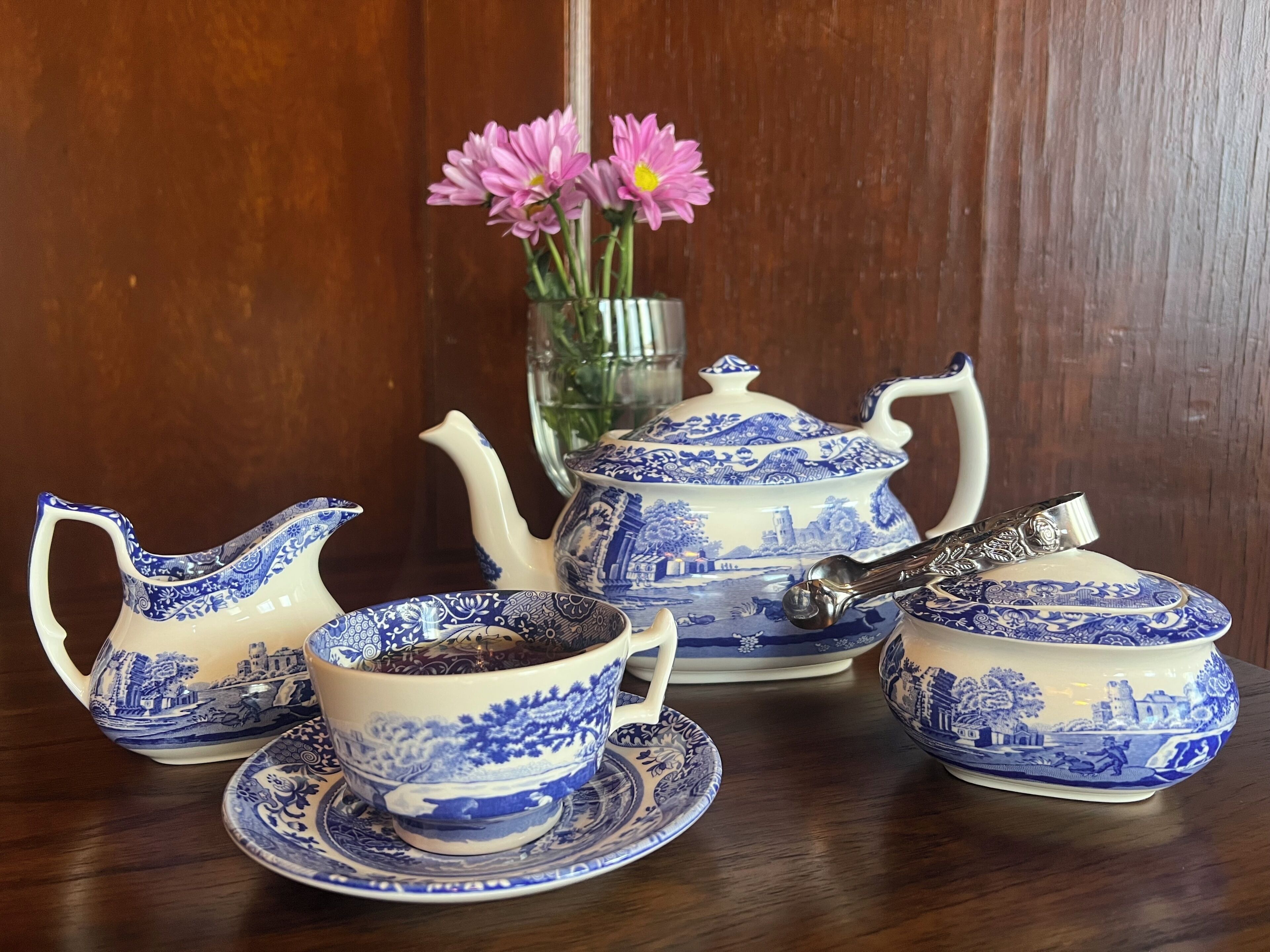 A tea set with a teapot, cups, and saucer, a sugar bowl, and a vase with flowers on a wooden table.
