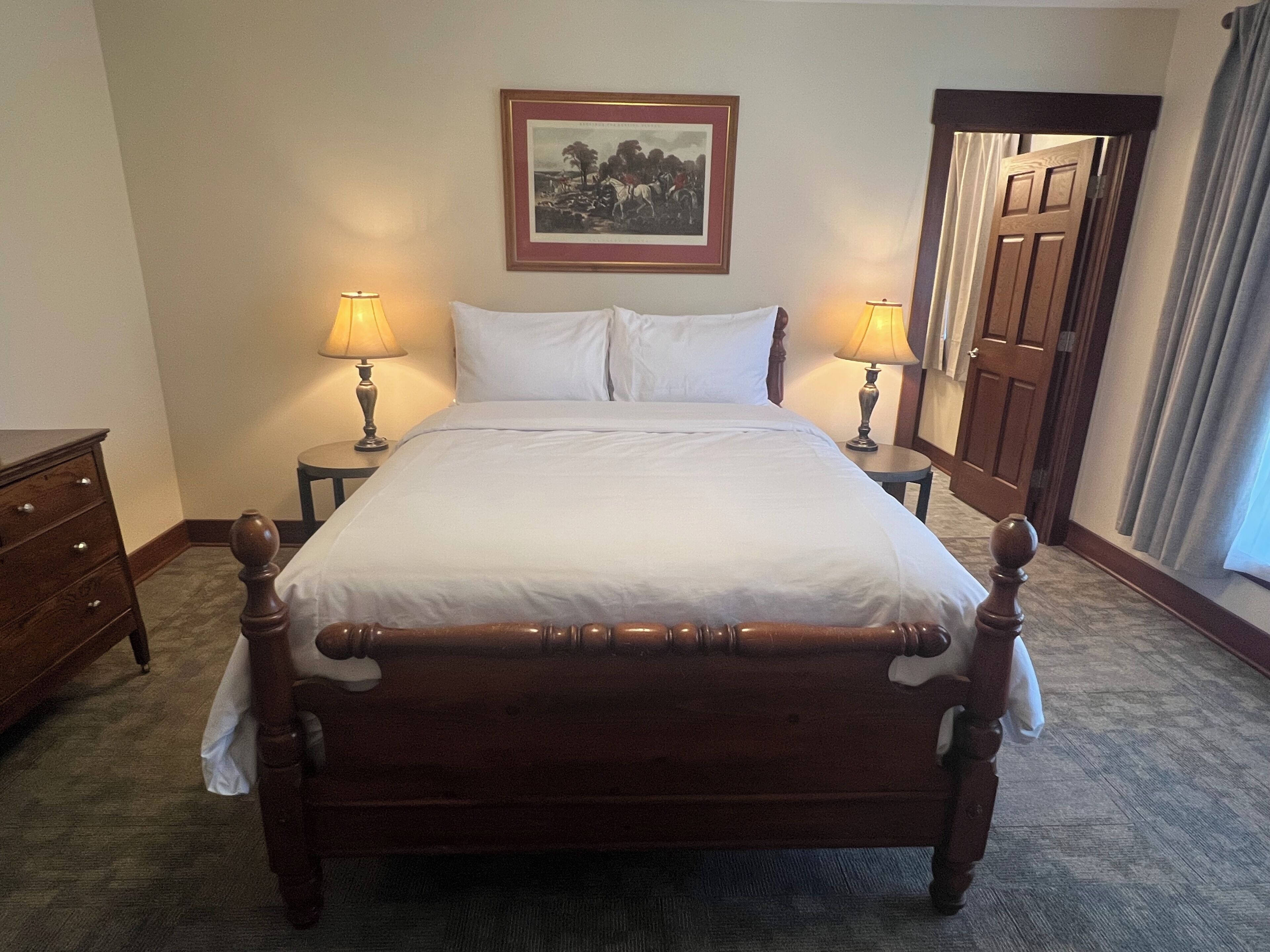 A hotel room with a large bed, two bedside tables with lamps, a wooden headboard, a framed picture on the wall, and a door with curtains.