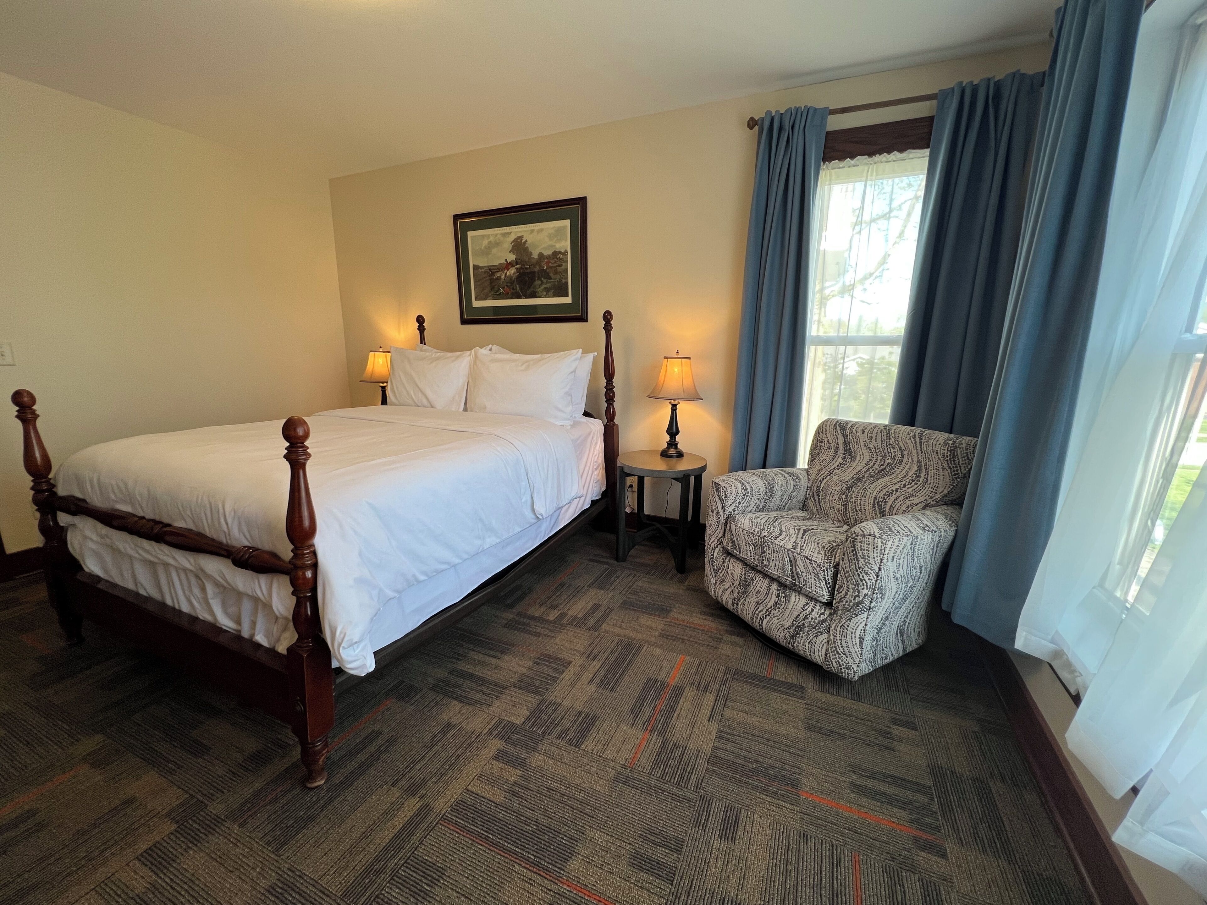 A hotel room with a large bed, a chair, a small table with a lamp, a framed picture on the wall, and a window with curtains.