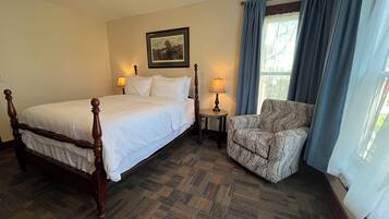 A hotel room with a large bed, a chair, a small table with a lamp, a framed picture on the wall, and a window with curtains.