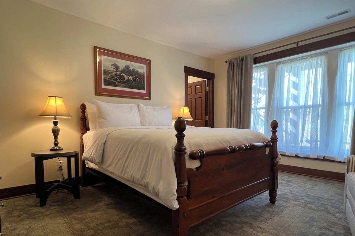 Una cama bien hecha en una habitación con un cuadro en la pared, una mesita con una lámpara y una ventana con cortinas.