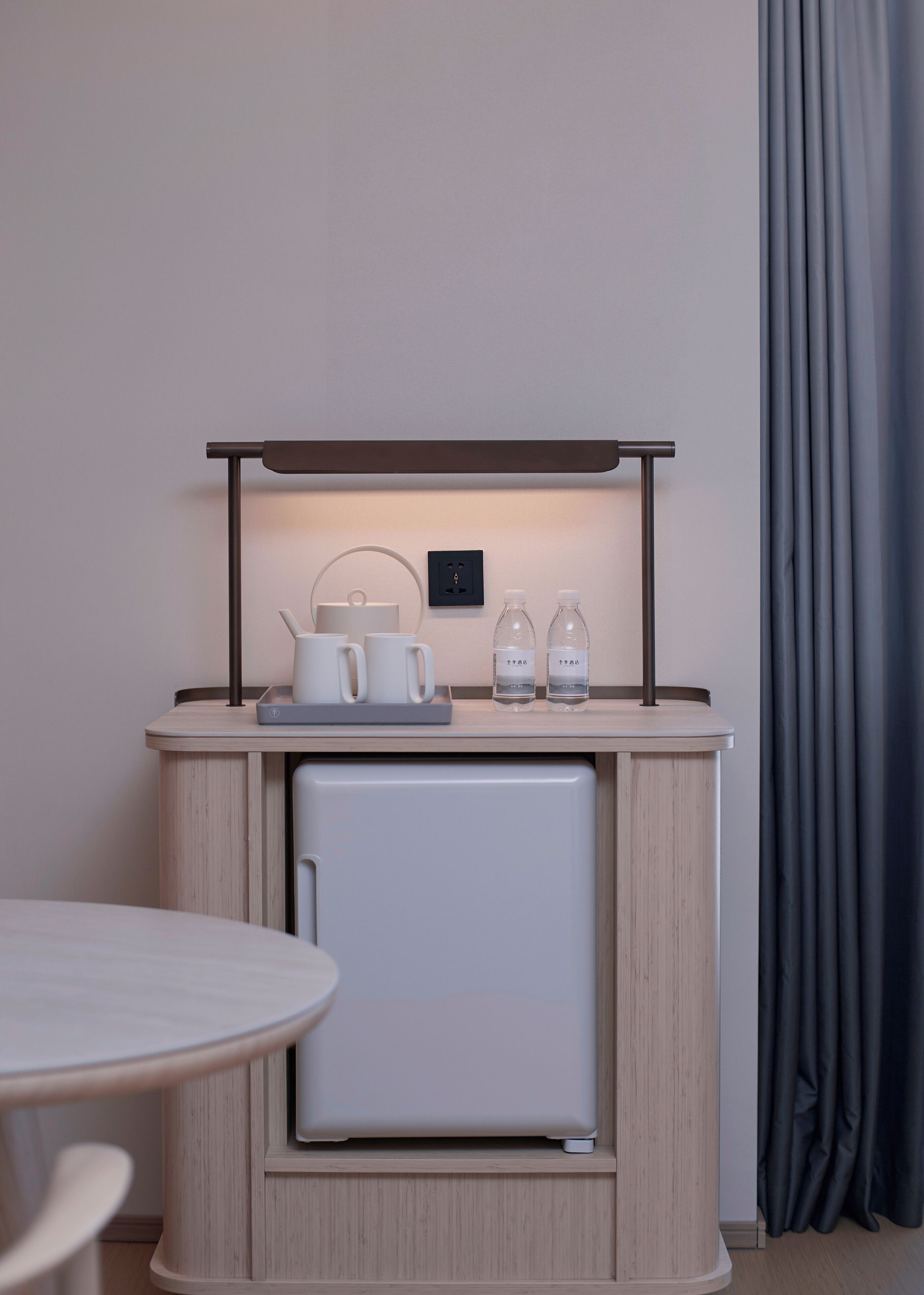 A compact kitchenette with a built-in fridge, a small table, and a countertop with a kettle, cups, and water bottles.