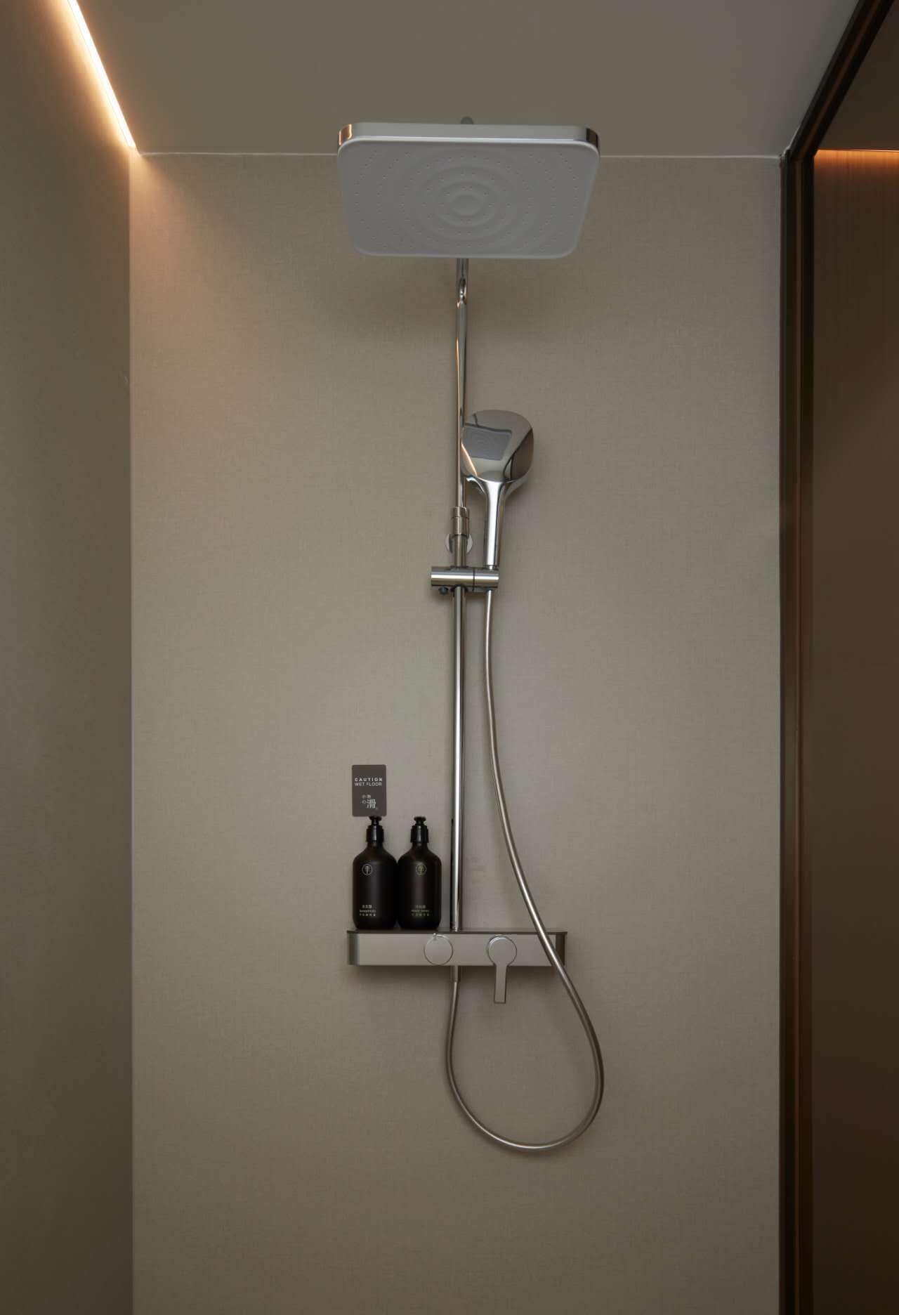 Een moderne douche met een handdouche en een vastgeplaatste douchekop, bevestigd aan een wand, met een plankje eronder waar twee flessen op staan.