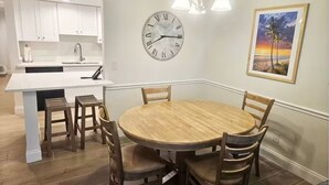 Un comedor con una mesa redonda de madera y cuatro sillas, un reloj de pared y un cuadro con una escena de playa.