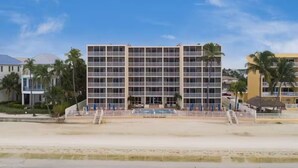 Un hotel de varios pisos con piscina frente a él, rodeado de palmeras y una playa.