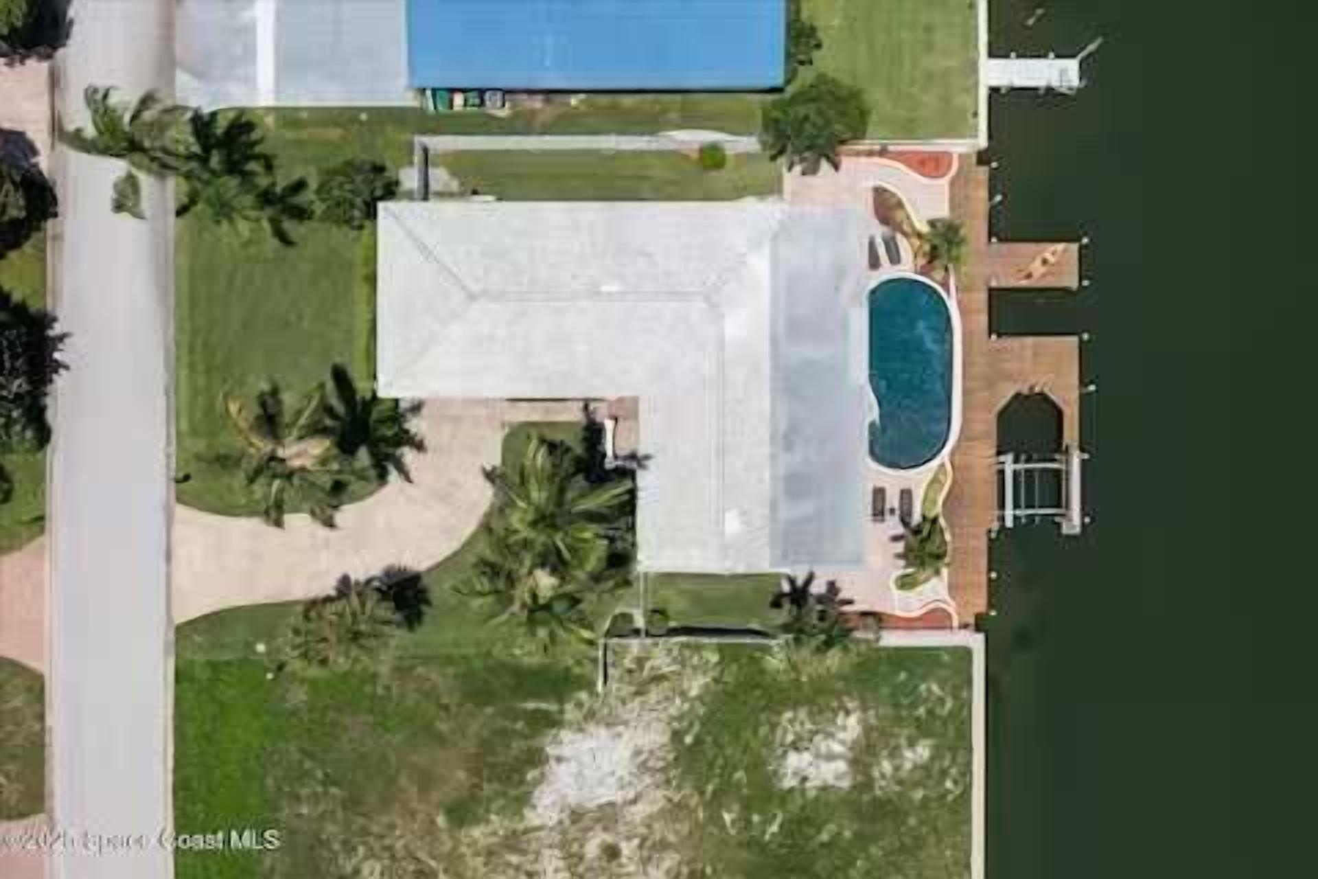 An aerial view of a property with a swimming pool, a covered patio, and a large lawn with palm trees.