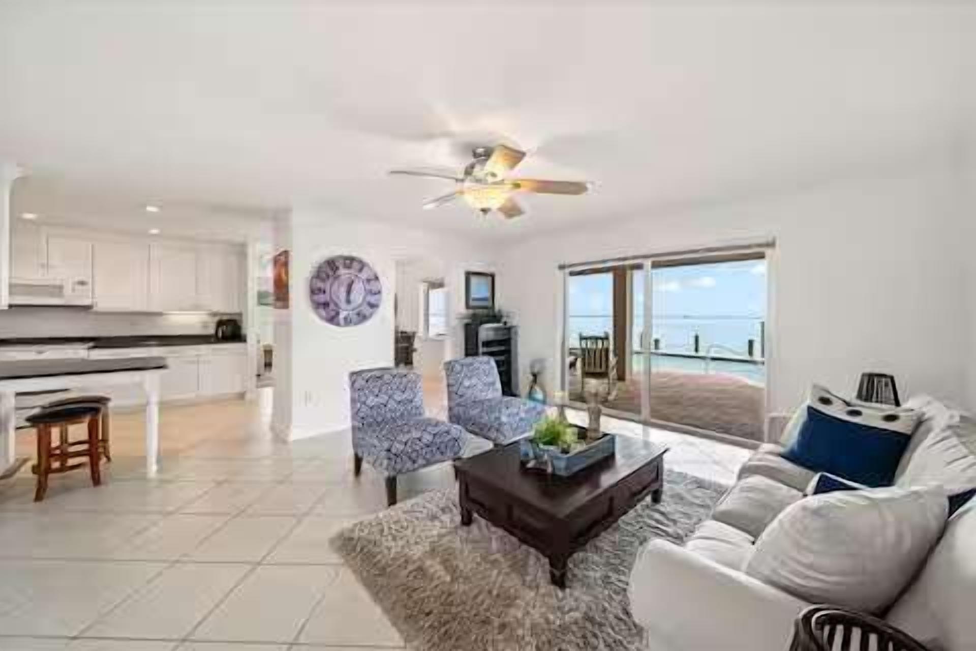 A spacious living area with a large window offering a view of the sea, a ceiling fan, and a modern kitchen in the background.