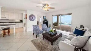 A spacious living area with a large window offering a view of the sea, a ceiling fan, and a modern kitchen in the background.