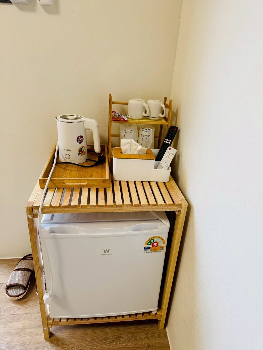 Eine kleine Küchenzeile mit Kaffeemaschine, kleinem Kühlschrank, einem Tablett mit Fernbedienung sowie einem Regal mit Tassen und Taschentuchbox.