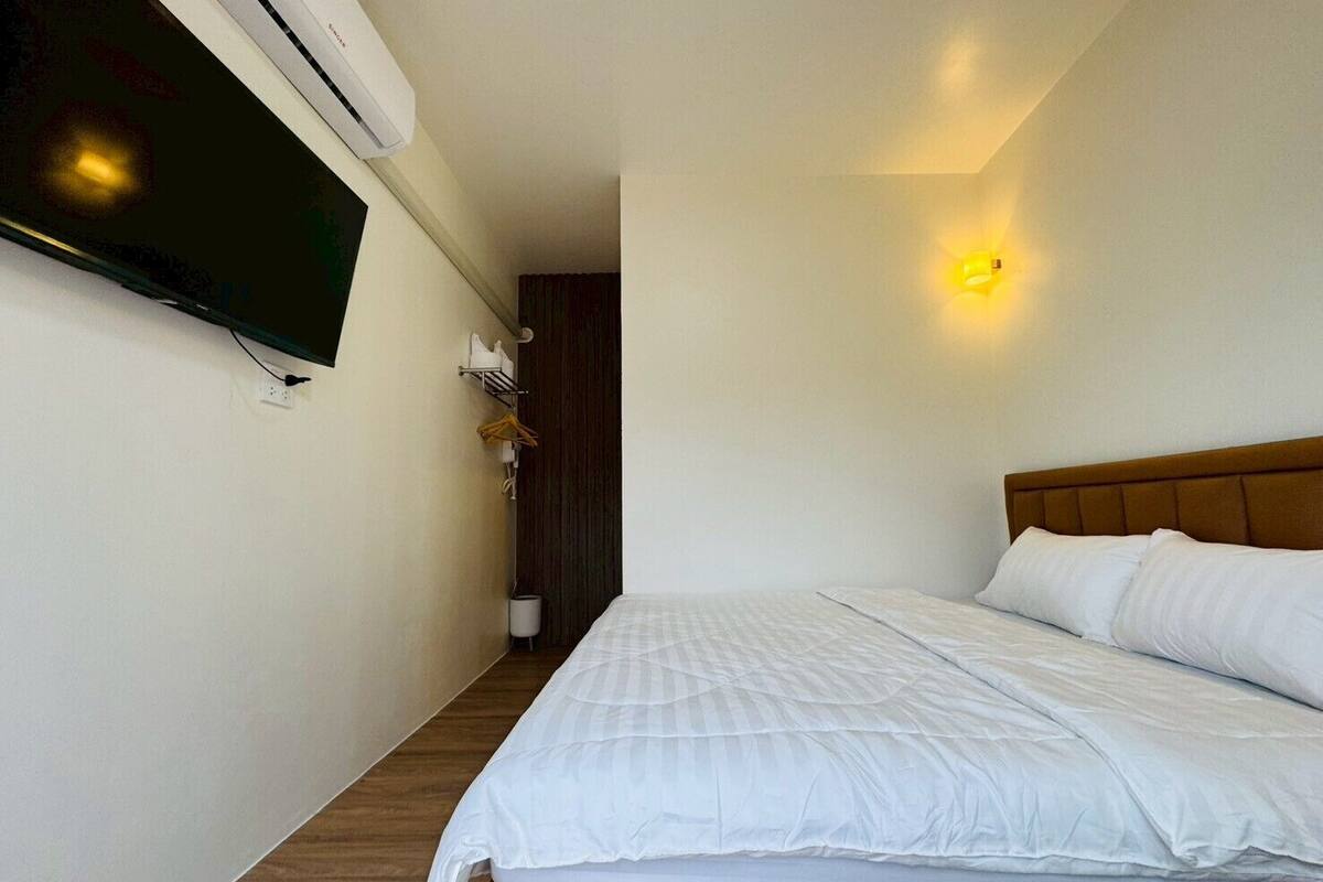 A hotel room with a bed, a flat-screen TV mounted on the wall, a bedside table with a lamp, and a wall-mounted air conditioner.