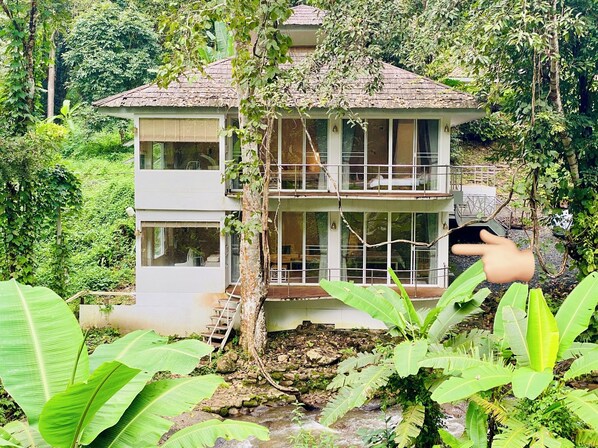 Un edificio de dos plantas con un balcón, rodeado de exuberante vegetación y un arroyo.