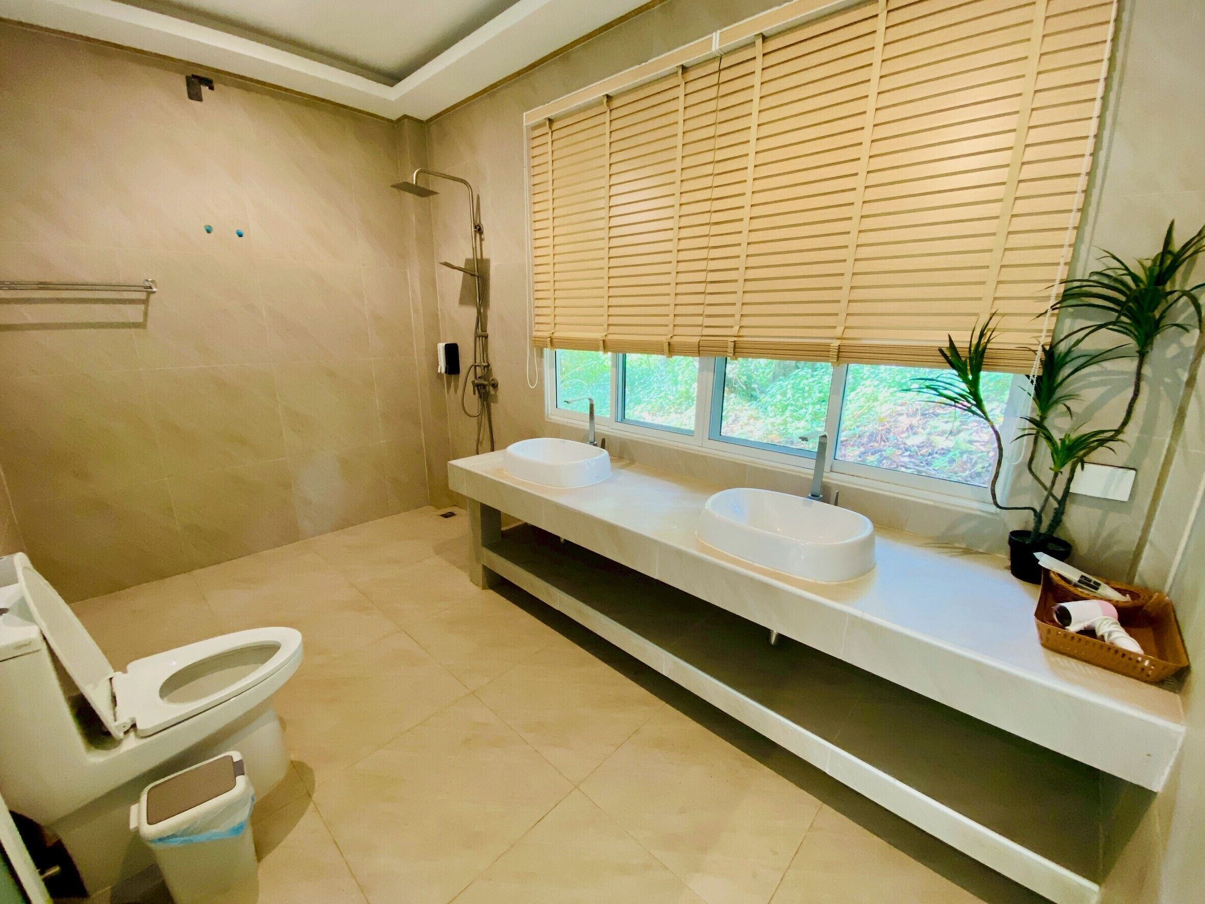 A bathroom with a toilet, two sinks, a shower, and a window with blinds.
