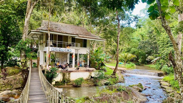 A wooden suspension bridge leading to a building labeled "Arb Pa Home & Cafe" with outdoor seating and a view of a flowing stream. - Arb Pa Home And Cafe At Mae On (Mae On)