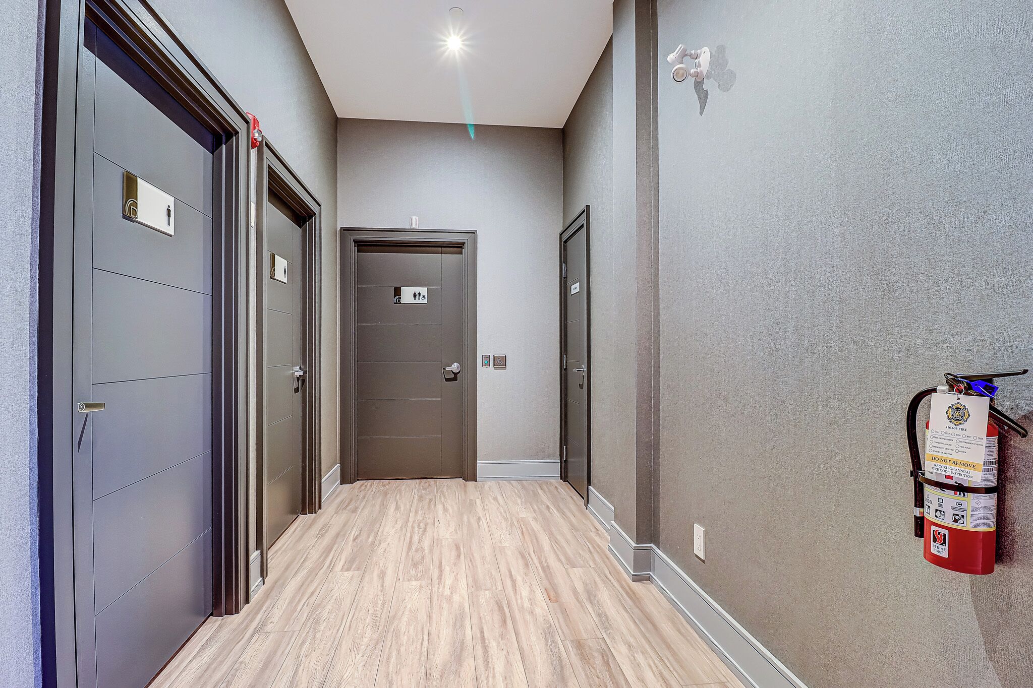 A hallway with grey doors, a fire extinguisher mounted on the wall, and a light fixture on the ceiling.