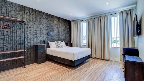A modern bedroom with a stone-patterned wall, a wooden floor, a bed with a headboard, a TV, and a window with curtains. - Dream Suites Near YYZ Airport (Mississauga)