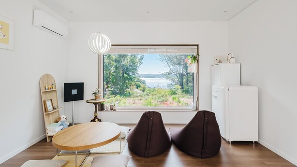 Una habitación moderna con un ventanal grande, una mesa redonda de madera, dos sillones tipo bean bag, un refrigerador blanco y un pequeño estante con objetos decorativos.
