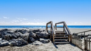 Una escalera de madera que conduce a una playa con rocas grandes al frente y el océano al fondo.