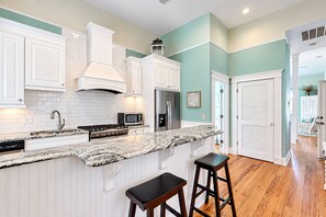 A kitchen with white cabinetry, a granite countertop, stainless steel appliances, and a wooden floor. - Charleston-style cottage w/ lovely decks, steps to the beach & pools (St. Simons Island)