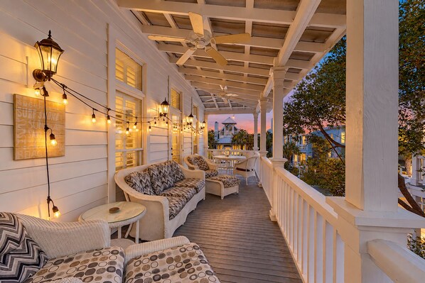 A covered porch with outdoor seating, string lights, and a view of a building in the background. - Charleston-style cottage w/ lovely decks, steps to the beach & pools (St. Simons Island)
