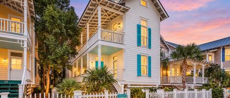 A row of white colonial-style buildings with balconies, a white picket fence, and a sign indicating rental availability.