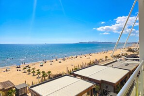 Vista da praia a partir de uma varanda, com céu claro, praia de areia e alguns guarda-sóis.