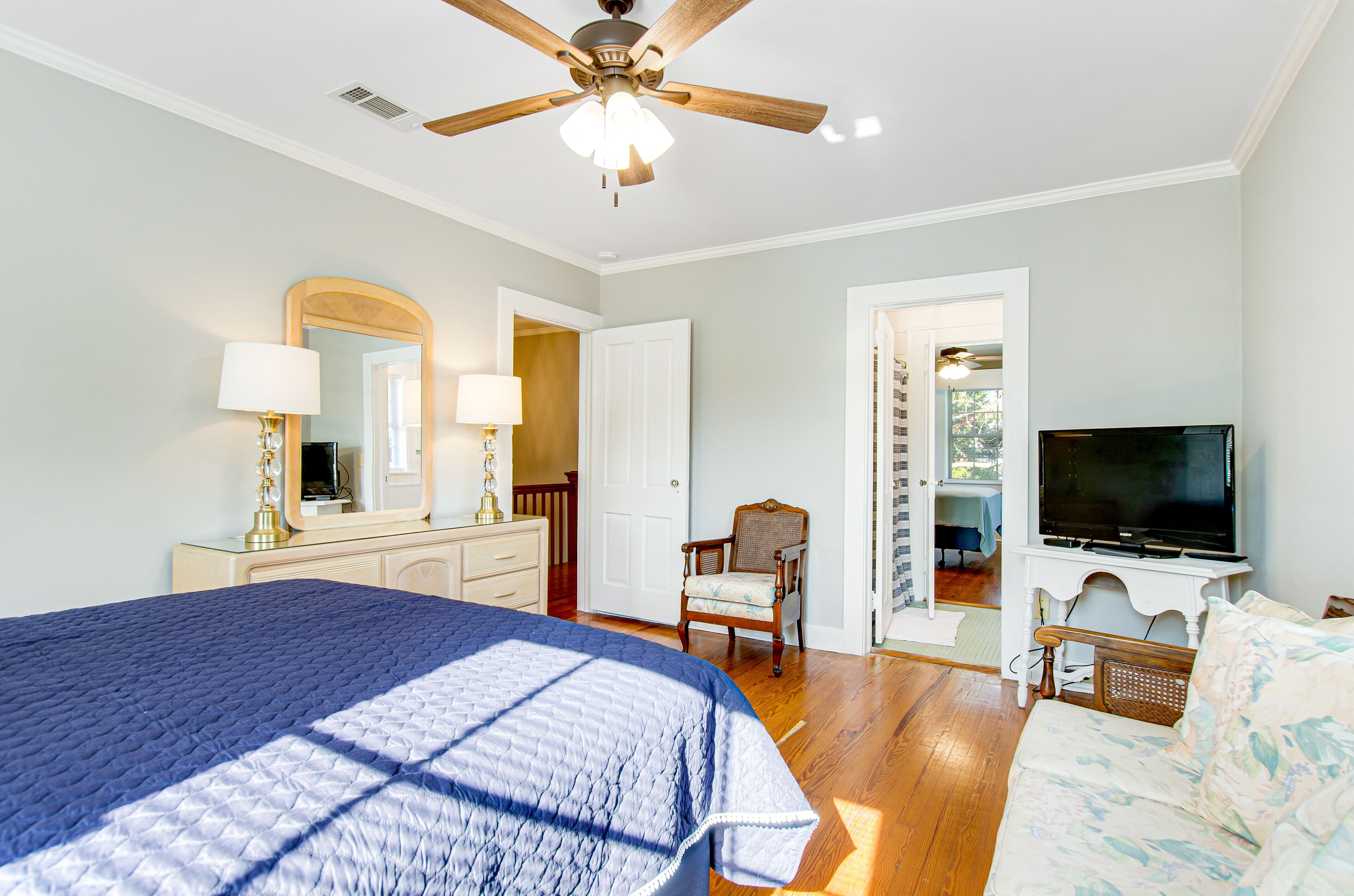 A bedroom with a bed, a television, a ceiling fan, a chair, and a door leading to another room.