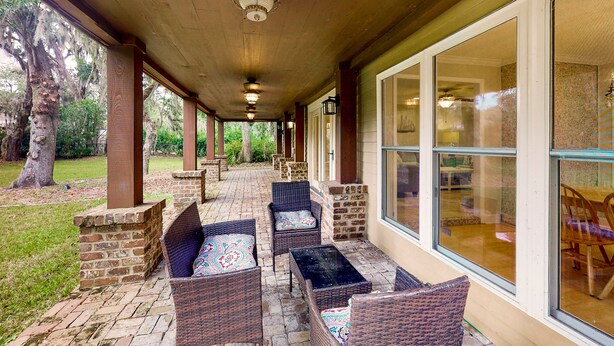 Un patio techado con mobiliario de mimbre y pilares de ladrillo, ventanas amplias y vistas al exterior.