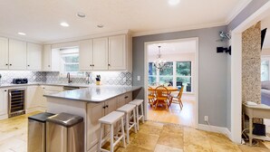 Una cocina moderna con gabinetes blancos, una isla grande con taburetes, horno empotrado y comedor con candelabro.