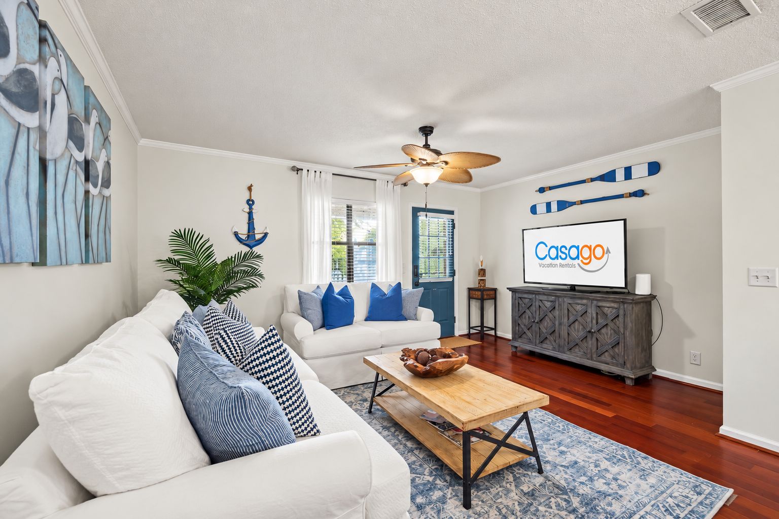 Ein Wohnzimmer mit einem weißen Sofa, blauen Kissen, einem Holztisch und einem Fernseher, der 'Casago Vacation Rentals' zeigt.
