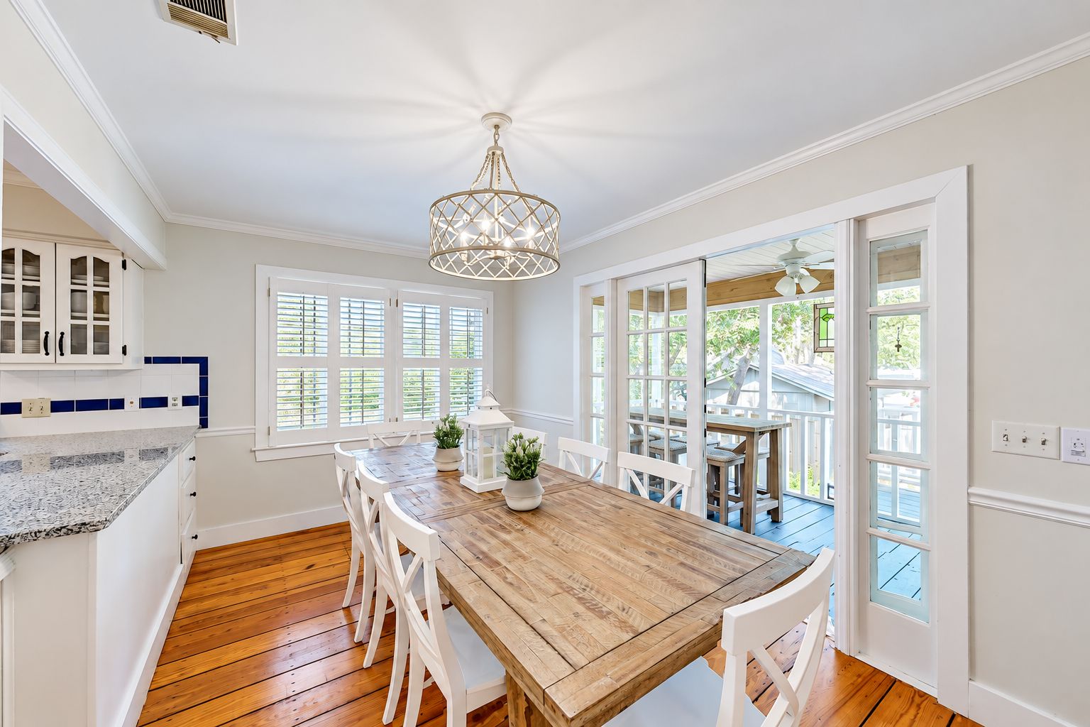 A dining area with a wooden table and white chairs, a chandelier, and large windows with shutters.