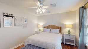 Une chambre à coucher comprenant un lit, un ventilateur de plafond, des tables de chevet avec des lampes et des œuvres d’art encadrées accrochées au mur.