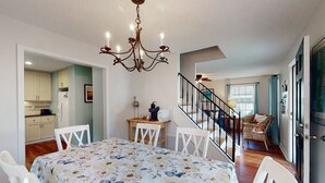 Un coin repas avec une nappe à motifs floraux, des chaises blanches, un lustre et une cuisine en arrière-plan.