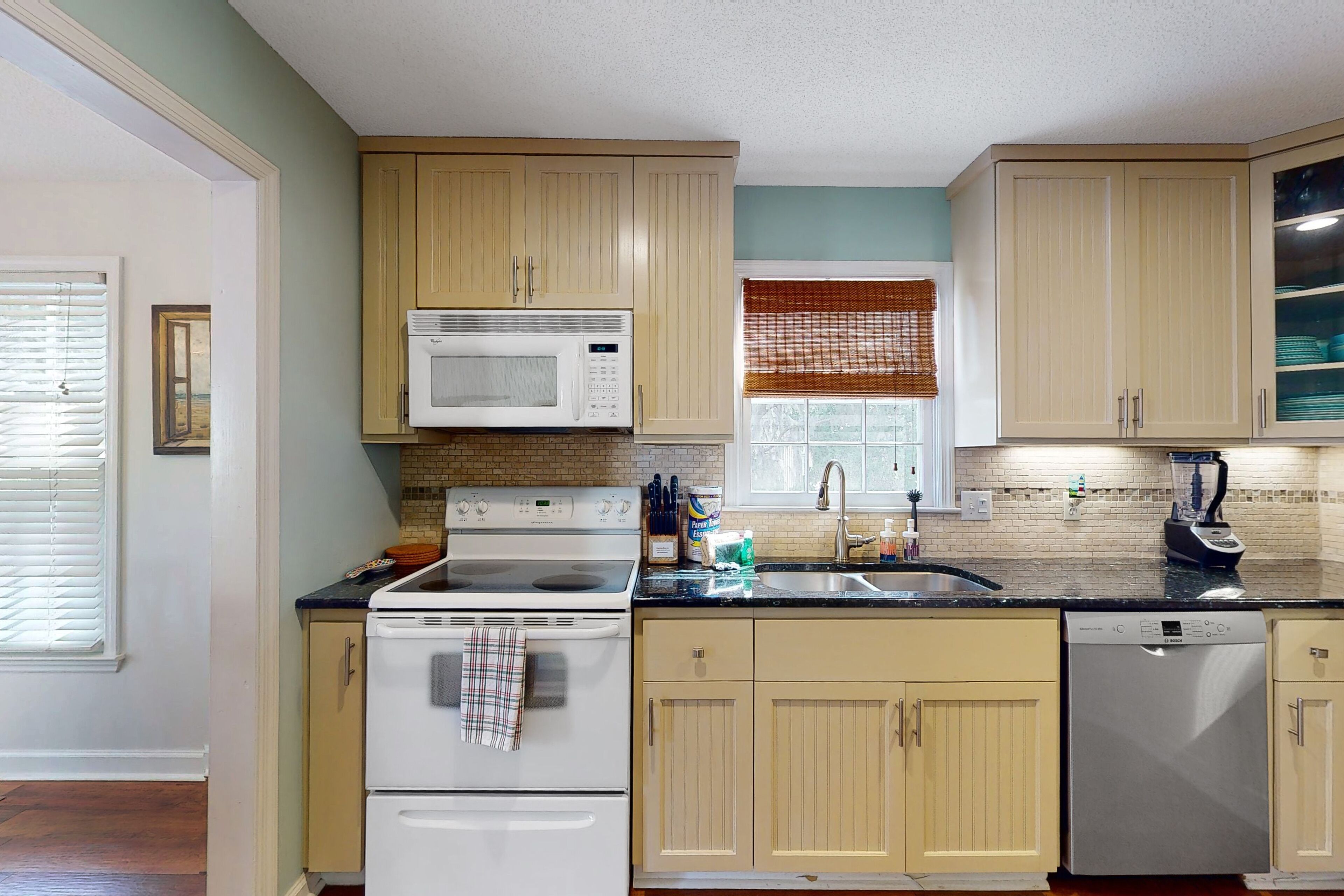 A kitchen with beige cabinets, a white stove, a microwave, a dishwasher, and a window with a brown blind.