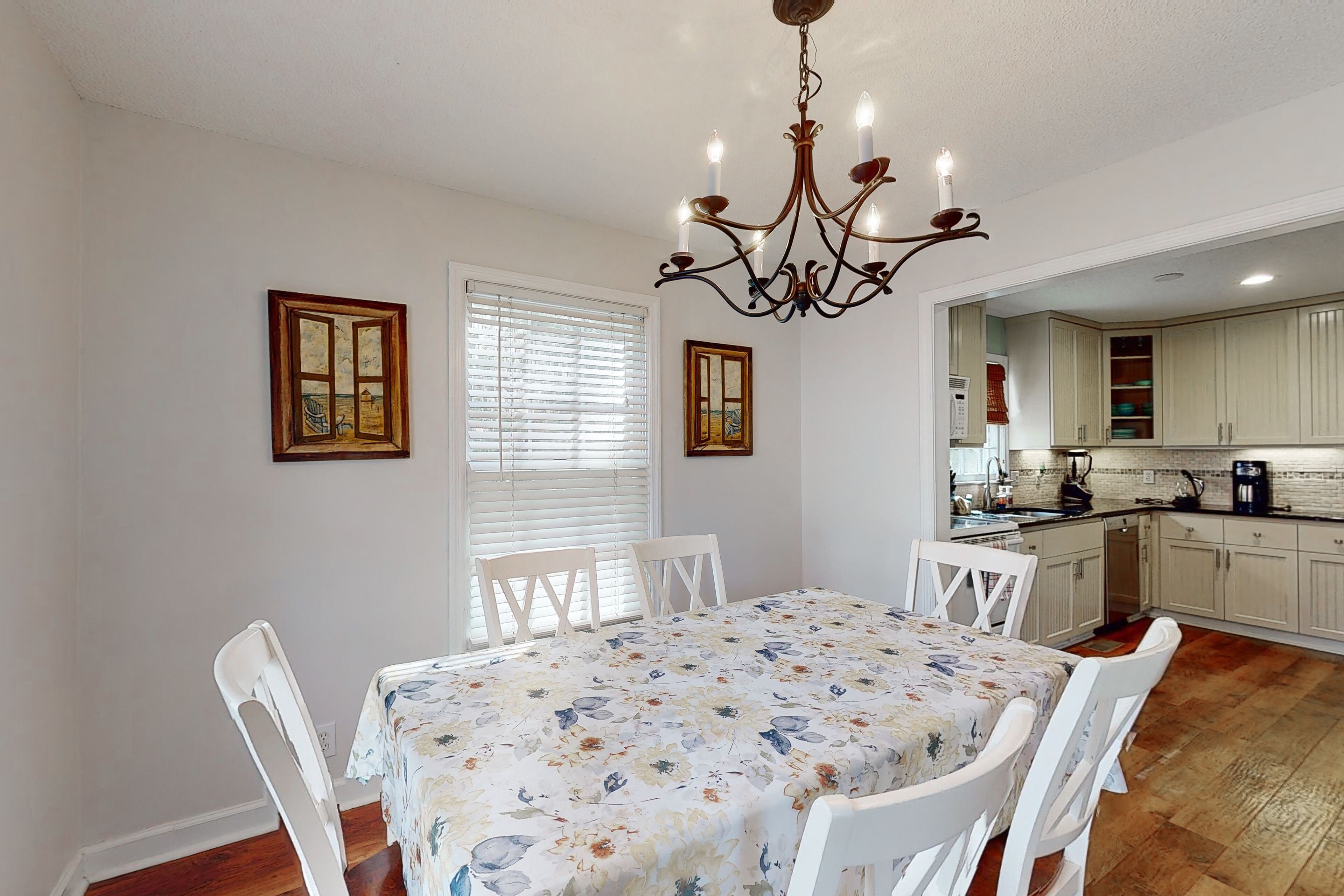 A dining area with a table set for four, white chairs, a chandelier, and a window with blinds.