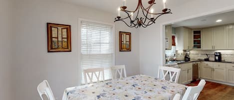 A dining area with a table set for four, white chairs, a chandelier, and a window with blinds.