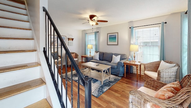 Un salon spacieux avec un escalier, un ventilateur de plafond, un canapé, une table basse et un fauteuil en osier.