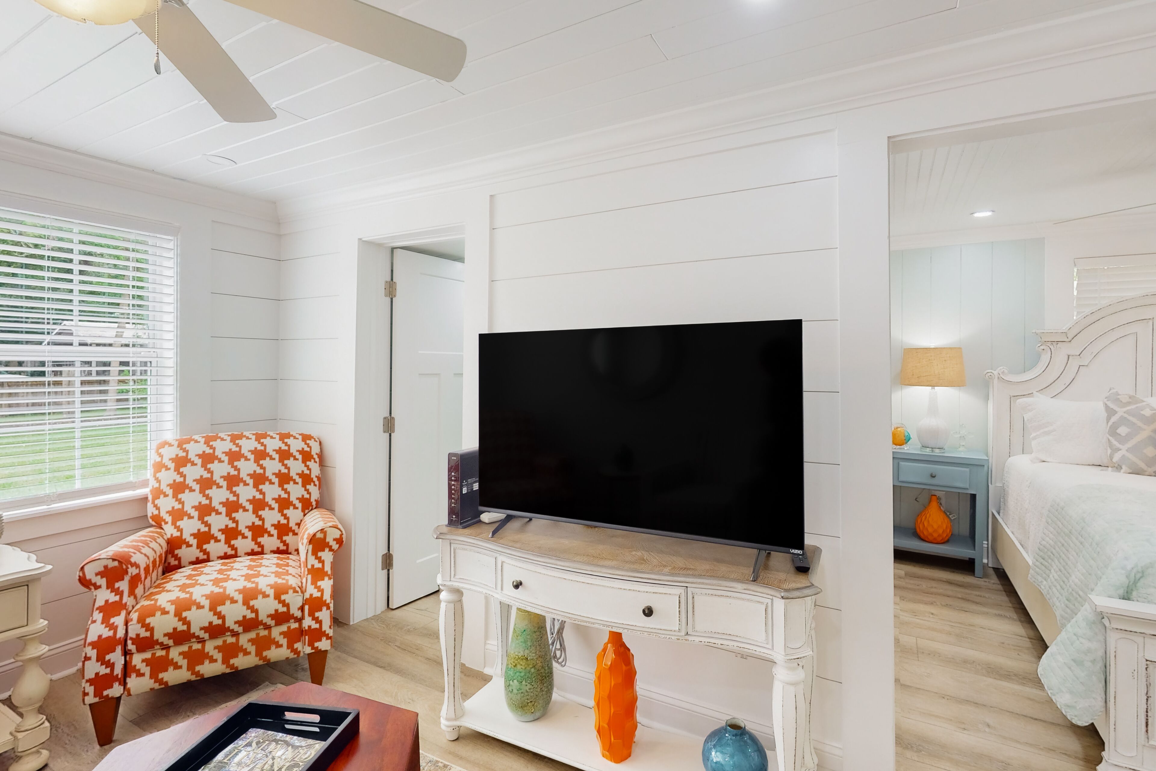 A room with a television on a white console, a houndstooth patterned armchair, and a bed with a nightstand.