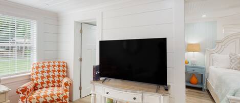 A room with a television on a white console, a houndstooth patterned armchair, and a bed with a nightstand.