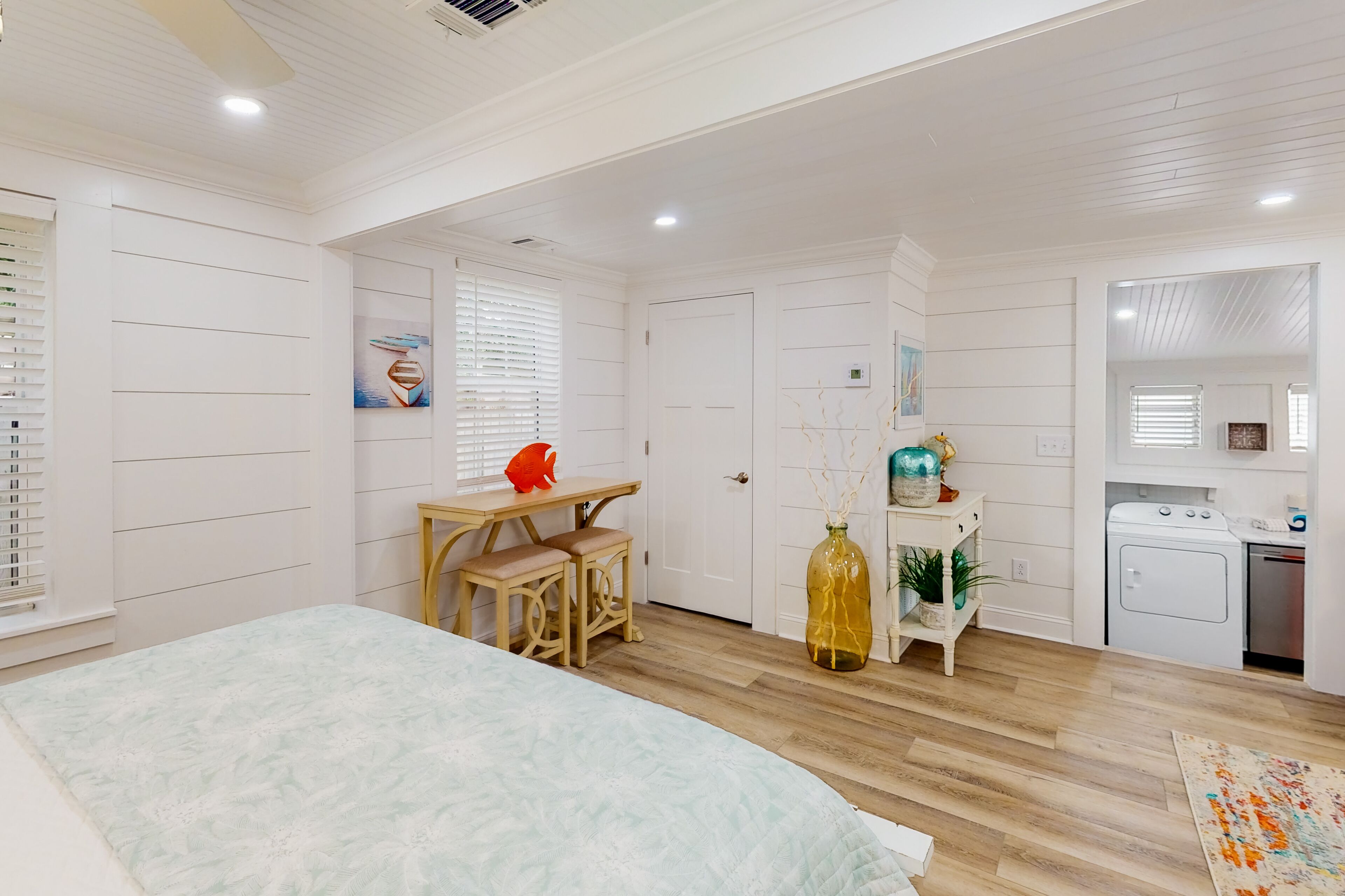 A bedroom with a bed, a desk, a chair, a window with blinds, a door, and a kitchenette.