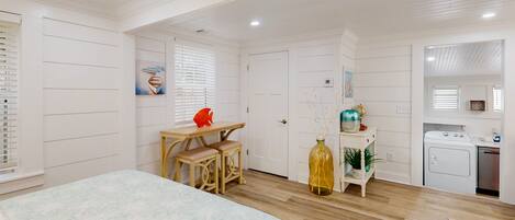 A bedroom with a bed, a desk, a chair, a window with blinds, a door, and a kitchenette.