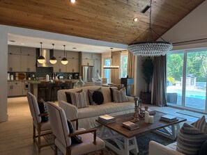 A modern living room with a wooden ceiling, a chandelier, a sofa, a coffee table, and a view of a swimming pool. - Lakefront home with private pool & outdoor space for entertaining (St. Simons Island)
