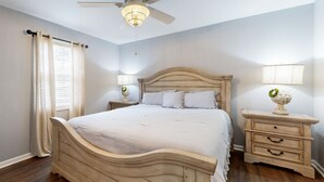 Une chambre à coucher avec un lit en bois, deux tables de chevet avec des lampes, un ventilateur de plafond et une fenêtre avec des rideaux.