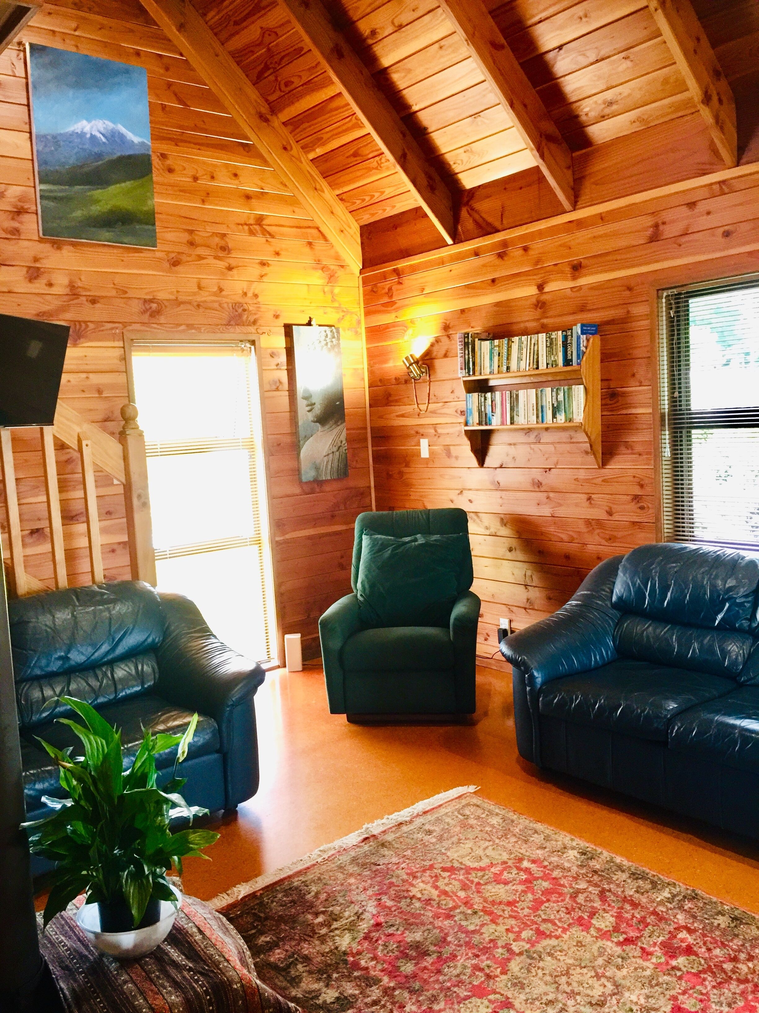 Un acogedor interior de cabaña de madera con un sillón verde, dos sofás de cuero azul, una estantería y un cuadro de una montaña.