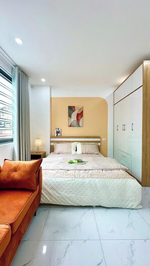 A modern hotel room with a large bed, a sofa, a nightstand, a lamp, a wardrobe, and a window with curtains. - 25 House 298 Ngoc Lam - Long Bien bridge (Long Biên)