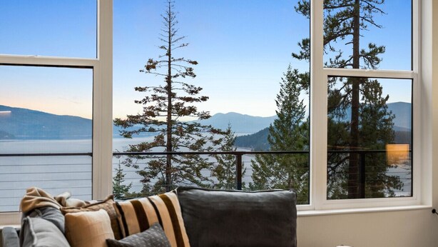 Une pièce avec un canapé gris, une petite table d’appoint en bois et une vue sur un lac et des montagnes, visible à travers de grandes fenêtres.