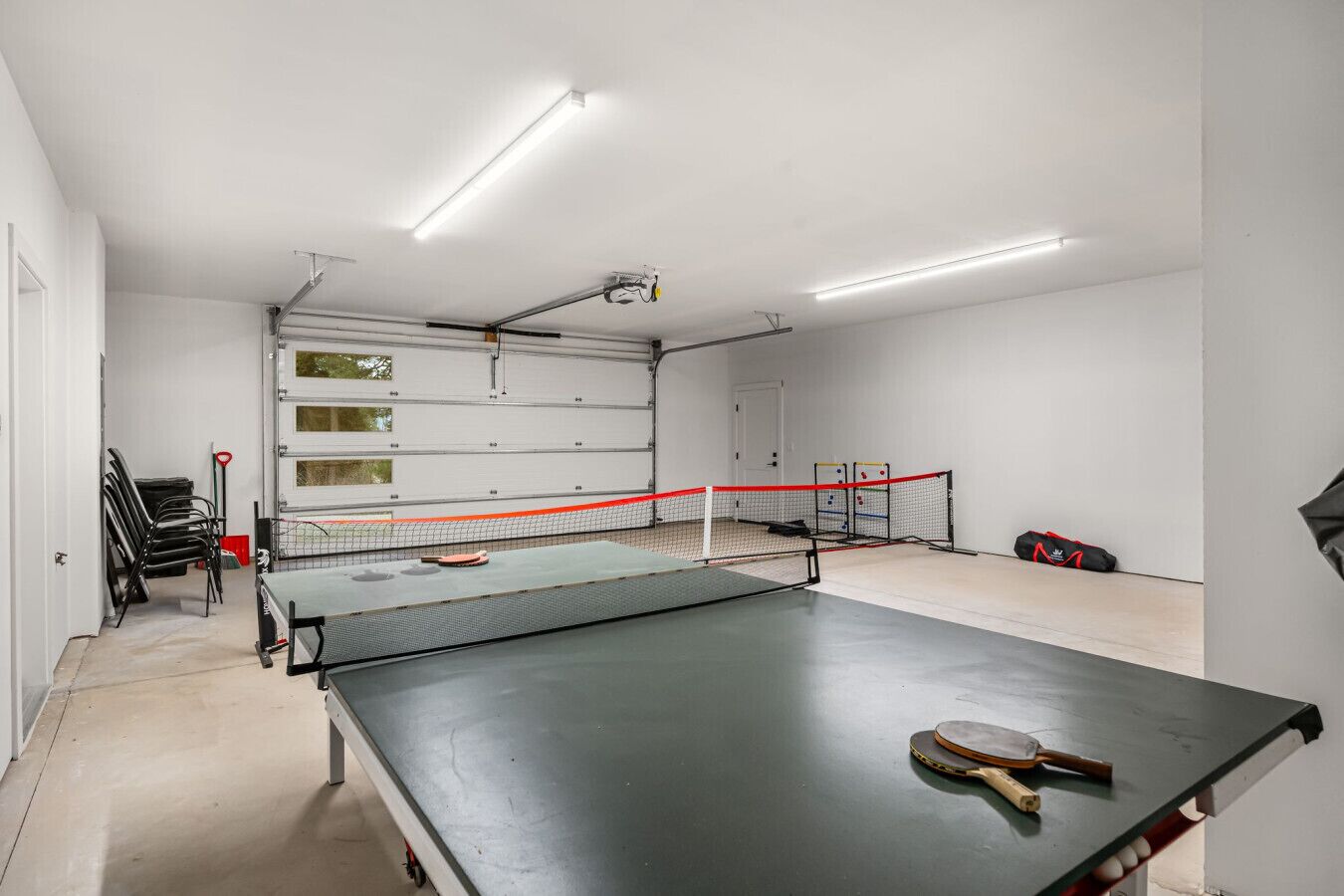 Une table de ping-pong avec un filet, dans une pièce aux murs blancs et dotée d’une porte de garage.